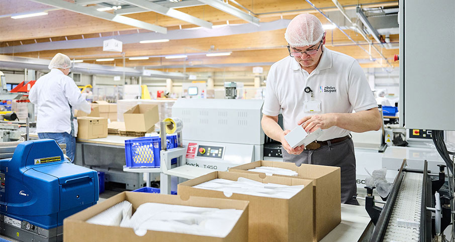 Un homme, porte un bonnet de protection, des lunettes, un polo avec un badge indiquant "Technical Support", et des gants emballe des articles dans une boite en carton qui se trouve sur un convoyeur